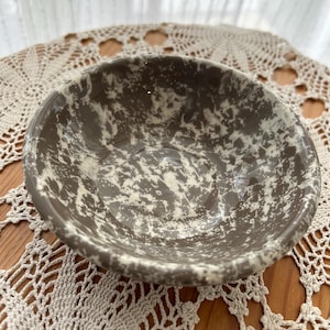 Puede incluir: Un cuenco redondo de cerámica con un patrón moteado y salpicado en colores topo y blanco roto. El cuenco tiene un acabado liso y brillante, con un interior profundo y cóncavo y un borde grueso y redondeado, mostrando un aspecto marmolado.
