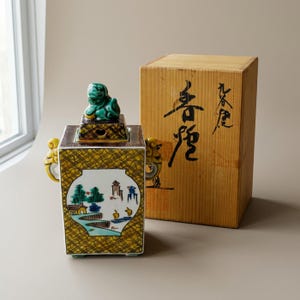 May include: A rectangular ceramic incense burner with a yellow and brown cross-hatched pattern, featuring a hand-painted landscape scene in a circular white panel, two side handles, and a lid topped with a green lion figure, beside a wooden box.