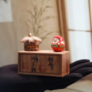 May include: A wooden box with Japanese characters holds two small figurines. One is a carved wooden mask with a straw roof, and the other is a red Daruma doll with white floral accents. The box is held in a black gloved hand.