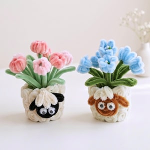 May include: Two decorative pots crafted from white textured material, each featuring a sheep face with googly eyes. One pot holds pink pipe cleaner flowers with green stems, while the other contains blue pipe cleaner flowers with green stems.