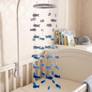 May include: A nursery mobile featuring a circular frame wrapped in light grey yarn, from which hang multiple strings of clear beads and small plush fish. The fish transition in colour from light grey at the top to deep blue at the bottom.