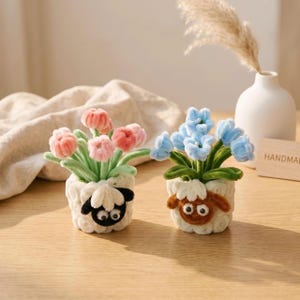 May include: Two small decorative pots shaped like sheep, one with a black face and one with a brown face, each holding a bouquet of fuzzy flowers. The left pot contains pink flowers with green stems, and the right pot contains light blue flowers.