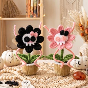 May include: Two pipe cleaner flowers in small tan pots. One flower has black petals with a pink bow, the other has pink petals with a black bow. Both feature white skull centers, green leaves, and pink stems shaped like arms, standing on a woven mat.