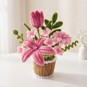 Peut inclure: Un bouquet de fleurs en cure-pipes roses et magenta dans un pot marron cannelé en forme de tasse avec une bordure festonnée blanche. L'arrangement comprend un grand lys central, une tulipe et des petites fleurs avec des tiges vertes.