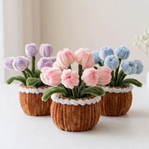May include: Three small potted arrangements featuring fuzzy pipe cleaner flowers in shades of lavender, light pink, and baby blue. Each plant has green stems and leaves, housed in a textured brown pot with a white braided trim along the top edge.