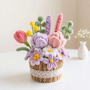 Puede incluir: Un arreglo decorativo de flores hechas con limpiapipas en tonos rosa, morado, amarillo y verde, dentro de una maceta marrón texturizada envuelta con una cinta de encaje blanco. El ramo presenta varias formas de pétalos, incluyendo tulipanes y margaritas.