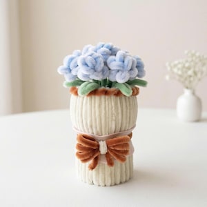 Pode incluir: Um buquê de flores de limpador de cachimbo azul-claro com folhas verdes está em um recipiente cilíndrico canelado de cor creme. O recipiente é decorado com um laço marrom felpudo e uma fita rosa claro enrolada ao redor do centro.