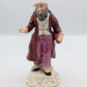 Puede incluir: Figura de cerámica de un hombre con un abrigo largo burdeos, chaleco blanco y pantalones morados. Lleva un sombrero blanco y tiene una larga barba. La figura está sobre una base texturizada con ribete dorado, sosteniendo un objeto rojo.