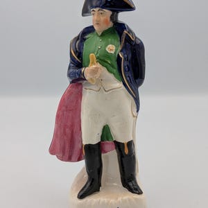 Puede incluir: Figura de cerámica de un personaje histórico, vistiendo un abrigo azul oscuro con ribetes dorados sobre una camisa verde y calzones blancos. La figura tiene un sombrero y botas negras, sosteniendo un objeto dorado. Una capa roja está drapeada.