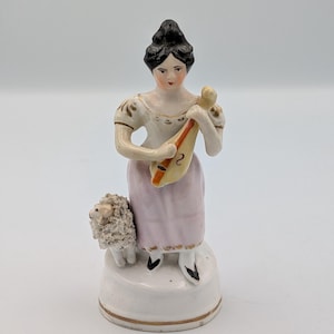 Puede incluir: Figura de porcelana de una mujer con un vestido blanco y rosa, sosteniendo un instrumento musical. Una pequeña oveja está a su lado. La figura tiene cabello negro y está sobre una base blanca con ribete dorado. El instrumento es amarillo y rojo.
