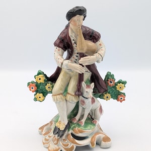 Puede incluir: Una figura de porcelana de un músico tocando la gaita, con una chaqueta de color rojo oscuro, calzones color crema y un gorro negro. Un pequeño perro blanco con manchas marrones se sienta a los pies del músico, flanqueado por follaje verde.