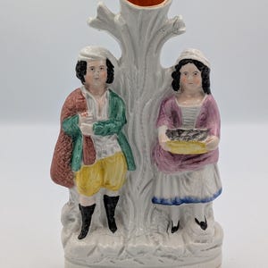 Puede incluir: Un jarrón de cerámica blanca con una estructura de árbol y una abertura central. Dos figuras con vestimenta de época se encuentran a ambos lados. El hombre lleva un abrigo verde, pantalones amarillos y botas negras. La mujer lleva una blusa rosa y sostiene una cesta.