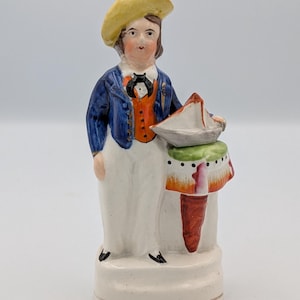 Puede incluir: Figura de cerámica vintage de una persona con chaqueta azul, chaleco naranja y pantalón blanco, con un sombrero amarillo. La figura sostiene un pequeño barco sobre una mesa con diseño verde y rojo. La base es blanquecina.