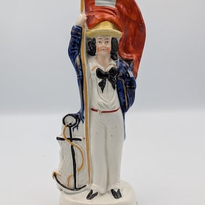 Puede incluir: Figura de cerámica vintage de un marinero sosteniendo una bandera roja con un cuadrado blanco. El marinero viste camisa blanca, pantalón y sombrero de paja, con chaqueta azul y pajarita negra. Un ancla está en la base.