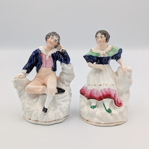 Puede incluir: Un par de figuras antiguas de porcelana que representan a un hombre y una mujer con ropa de época. El hombre lleva una chaqueta azul, un chaleco rosa y pantalones color melocotón. La mujer viste un vestido blanco con un detalle rojo y rosa. Ambas figuras están sobre bases blancas.