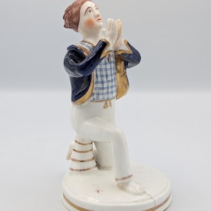 Puede incluir: Figura de porcelana de una figura arrodillada con las manos juntas en oración, mirando hacia arriba. La figura viste una chaqueta azul con ribetes dorados sobre un chaleco a cuadros, pantalones blancos y tiene el pelo castaño. La base es blanca con detalles dorados.
