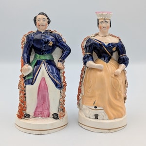 Puede incluir: Dos figuras antiguas de porcelana de un hombre y una mujer. El hombre viste un abrigo azul con detalles dorados, una faja verde y una prenda interior rosa. La mujer lleva un vestido azul con falda amarilla y sombrero rosa. Ambos tienen adornos ornamentados.