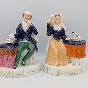 Puede incluir: Dos figuras de cerámica que representan a un hombre y una mujer sentados. El hombre lleva un abrigo azul y pantalones verdes, la mujer un vestido azul y beige. Cada figura tiene una pequeña mesa con una taza y un platillo.