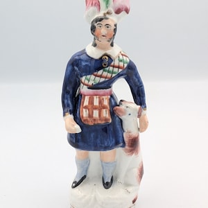 Może przedstawiać: Figurka ceramiczna w stylu vintage przedstawiająca mężczyznę w niebieskim płaszczu i kolorowym kapeluszu, trzymającego psa. Figurka ma białą podstawę i szczegółowy wzór z czerwonymi, zielonymi i białymi akcentami.