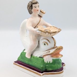 Könnte beinhalten: Porzellanfigur, die einen Cherub darstellt, der auf einem Delphin reitet. Der Cherub hält eine muschelförmige Schale. Der Sockel ist grün und lila mit goldenen Akzenten. Der Delphin hat goldene Verzierungen und einen rosa Mund.