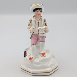 Puede incluir: Figura de porcelana de una figura sentada con atuendo del siglo XVIII. La figura lleva una chaqueta blanca con ribetes dorados, pantalones rosas y un sombrero blanco con ala dorada. Sobre una base blanca con detalles dorados, sosteniendo una pequeña taza.
