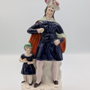 Puede incluir: Una figura de cerámica que representa a dos figuras, un adulto y un niño, con atuendo azul oscuro. La figura adulta lleva un sombrero con una pluma rosa y una capa roja y verde. La base de la figura está inscrita con las palabras "William Tell."