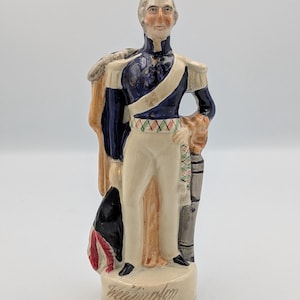 Puede incluir: Una figura de cerámica de un hombre con uniforme militar. La figura lleva una chaqueta azul marino con detalles dorados, pantalones color crema y una capa beige. La base de la figura está inscrita con el nombre "Wellington".