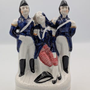 Puede incluir: Una figura de cerámica que representa a tres figuras con uniforme militar histórico. Dos soldados con abrigos azules y sombreros negros flanquean una figura central con un atuendo rojo y azul. La base es blanca con una raya roja.
