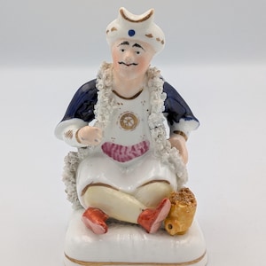 Puede incluir: Figura de porcelana de una figura sentada con turbante blanco y atuendo azul y blanco. La figura tiene un delantal blanco, una faja rosa y zapatos rojos. Una pequeña bolsa de color canela está a su lado.