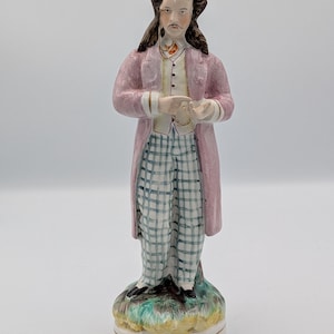Puede incluir: Figura de cerámica de un hombre con abrigo rosa y pantalones a cuadros. La figura tiene pelo largo y oscuro y bigote. La base es circular con detalles verdes y amarillos.