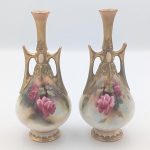 May include: A pair of porcelain vases featuring a bulbous base with hand-painted pink roses and green leaves. The vases have a narrow neck and two ornate gold-toned handles extending from the neck to the shoulder, with a gold-trimmed base.