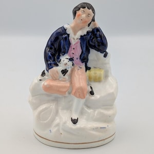 Op de afbeelding: Een porseleinen beeldje dat een zittende figuur met een kleine hond afbeeldt. De figuur draagt een donkerblauwe jas, een roze vest en perzikkleurige broek. De hond is wit met zwarte vlekken. Het beeldje staat op een witte basis.