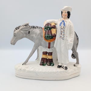 Pode incluir: Uma estatueta de cerâmica representando uma pessoa com casaco e chapéu brancos ao lado de um burro cinzento. O burro transporta uma alforje com fardos verdes e castanhos, e dois fardos adicionais repousam na base, que apresenta o texto DE BRUYN.