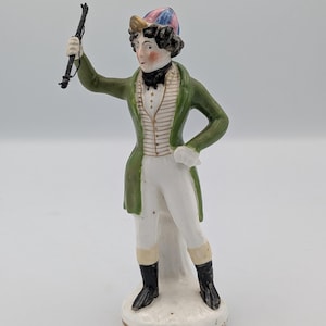 Puede incluir: Figura de porcelana vintage de un hombre con abrigo verde, chaleco blanco y calzones. La figura lleva un sombrero con detalles rosas, azules y amarillos y sostiene un látigo. La base es blanca con ribete dorado.