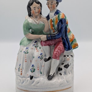 Puede incluir: Figura de cerámica que representa a una pareja con vestimenta del siglo XVIII. La mujer lleva un vestido verde, y el hombre un abrigo azul y un chaleco estampado. Un perro pequeño está a sus pies. La base es blanca con ribete dorado.
