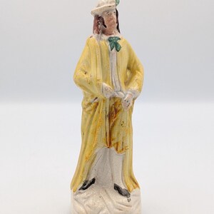 Könnte beinhalten: Vintage Keramikfigur einer Person in gelbem Mantel, weißem Hemd und Hut. Die Figur hat eine grüne Fliege und schwarze Schuhe. Die Figur steht auf einem weißen Sockel mit rissiger Oberfläche.