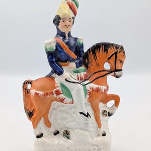 Puede incluir: Figura de cerámica pintada a mano que representa a un soldado a caballo. El soldado viste un uniforme azul con una faja roja y un sombrero alto y colorido. El caballo es naranja con detalles negros, y la base es blanca.