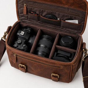Puede incluir: Una bolsa de cámara de cuero marrón con la parte superior abierta, que presenta un interior marrón oscuro con divisores acolchados ajustables. La bolsa contiene un cuerpo de cámara negro, dos lentes y varios accesorios en los bolsillos.
