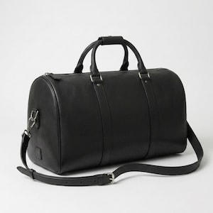 May include: A black duffel bag featuring a smooth finish, two top handles with a central grip, and a detachable, adjustable shoulder strap. The bag includes silver-toned metal hardware, a top zipper closure, and vertical strap detailing.