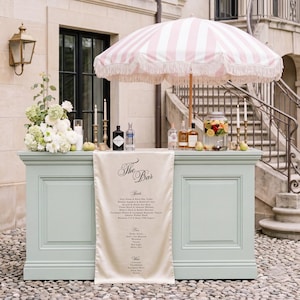 May include: A light green bar counter with a cream-coloured runner featuring the text The Bar and a list of drinks. Above the counter is a pink and white striped umbrella with fringe, accompanied by floral arrangements, candles, and bottles.