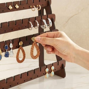 May include: A dark brown wooden earring display stand with multiple rows of slots for hanging earrings. Various earrings are displayed, including gold hoops, silver hoops, and woven teardrop earrings. A hand is placing an earring on the stand.
