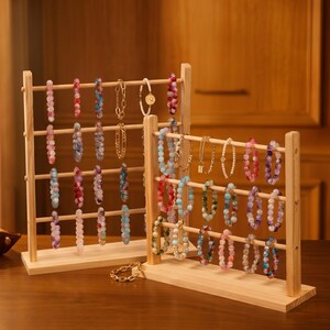May include: Two wooden bracelet display stands showcasing various beaded bracelets and gold chain bracelets. The bracelets feature a variety of colors, including pink, blue, and green. The stands are made of light-colored wood.