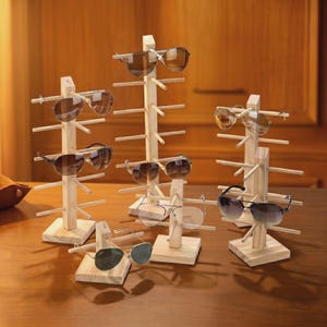 Peut inclure: Plusieurs présentoirs à lunettes de soleil en bois sont disposés sur une surface en bois. Chaque présentoir a une base carrée et une structure verticale avec des chevilles horizontales pour tenir les lunettes de soleil. Diverses lunettes de soleil sont exposées.