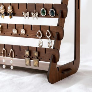 May include: A dark brown wooden earring display stand with multiple rows of slots for hanging earrings. Various earrings are displayed, including studs, hoops, and dangling styles. The stand has a modern, minimalist design, perfect for organizing and showcasing jewelry.