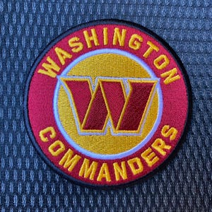 May include: A circular embroidered patch featuring the Washington Commanders logo. The design includes a large burgundy letter W centered on a gold circle, surrounded by a burgundy border with the text WASHINGTON COMMANDERS in yellow.