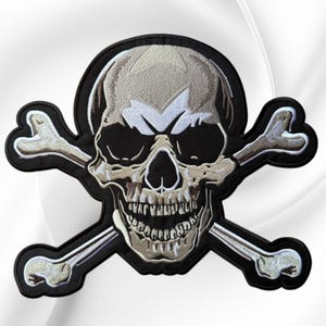 May include: An embroidered patch featuring a central human skull with a wide, grinning jaw, rendered in shades of beige, white, and black. Two crossed bones are positioned behind the skull, all outlined in a thick black border on fabric.