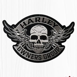 May include: A black and silver embroidered patch featuring a central skull design with wings extending to the sides. The text HARLEY is arched above the skull, and OWNERS GROUP is arched below, all set against a dark background with a border.