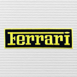 May include: Embroidered patch featuring the word "Ferrari" in yellow block letters on a black rectangular background. The patch has a yellow border and is likely designed to be sewn or ironed onto fabric.