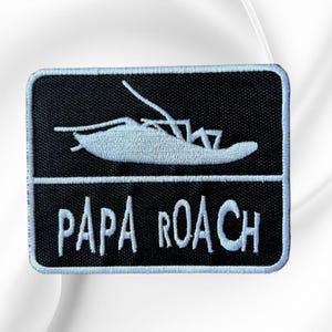 May include: A rectangular embroidered patch with a black background and a white border. The top section features a white graphic of a cockroach. The bottom section contains the white text "PAPA ROACH" in a stylized, hand-drawn font.