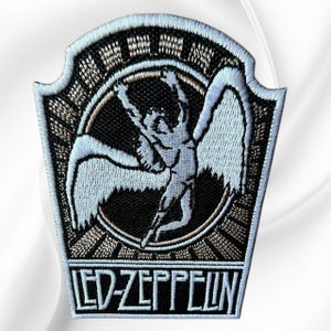 May include: A shield-shaped embroidered patch featuring a black background with a white winged figure centered within a circular border. The design includes a sunburst pattern above the circle and the text LED-ZEPPELIN in white at the bottom.